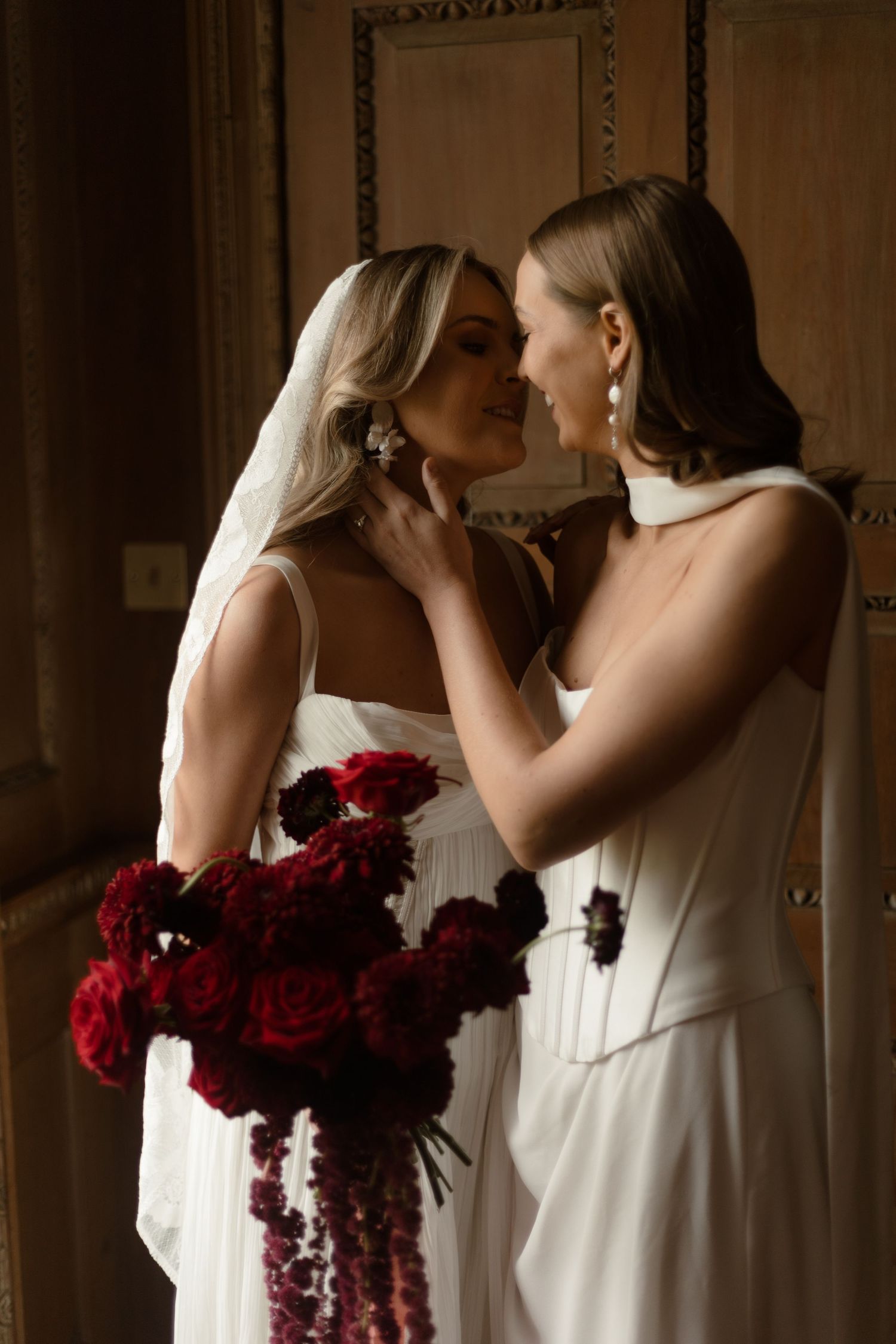 Two brides in white dresses share an intimate moment with a dramatic bouquet of deep red roses.