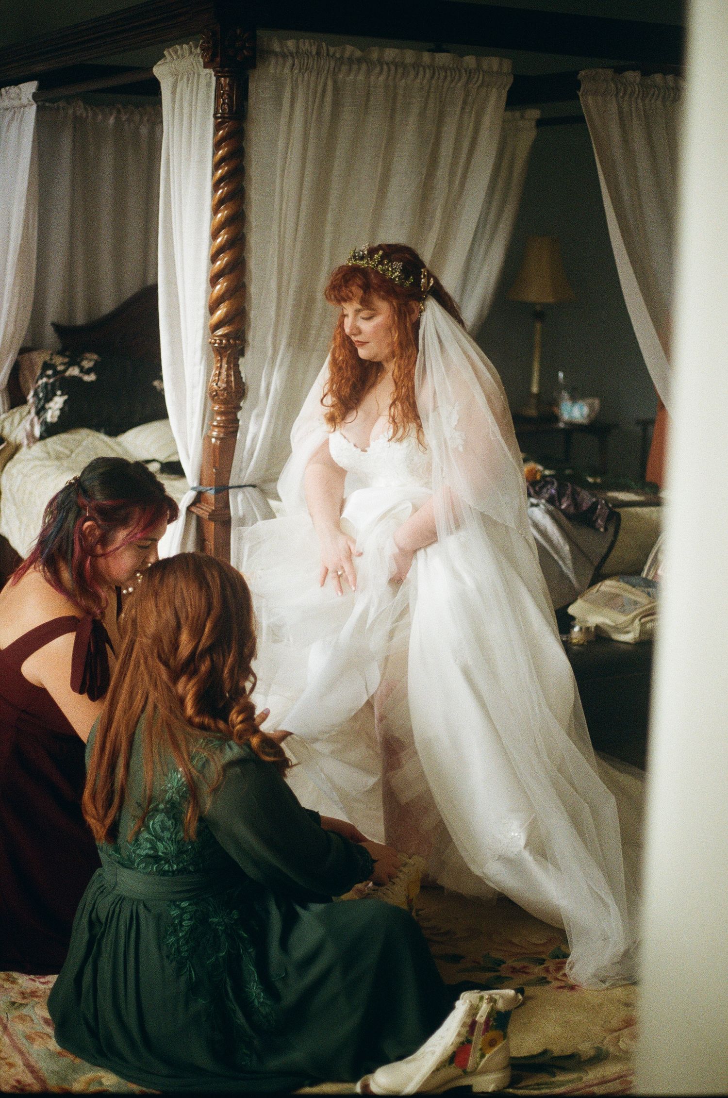 A bride in a flowing white wedding dress stands by a four-poster bed while attendants adjust her gown in a romantic setting.