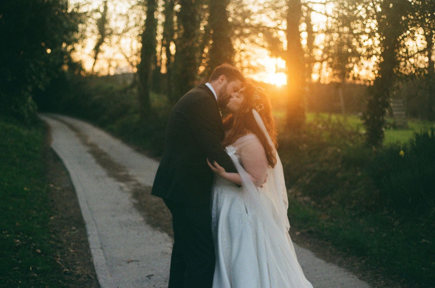 A romantic wedding couple embraces on a sunlit path through the woods during golden hour sunset.