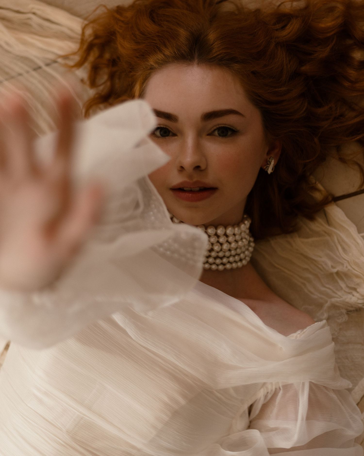 A fashion portrait in sepia tones shows a model in pearls reclining against flowing fabric with soft lighting.