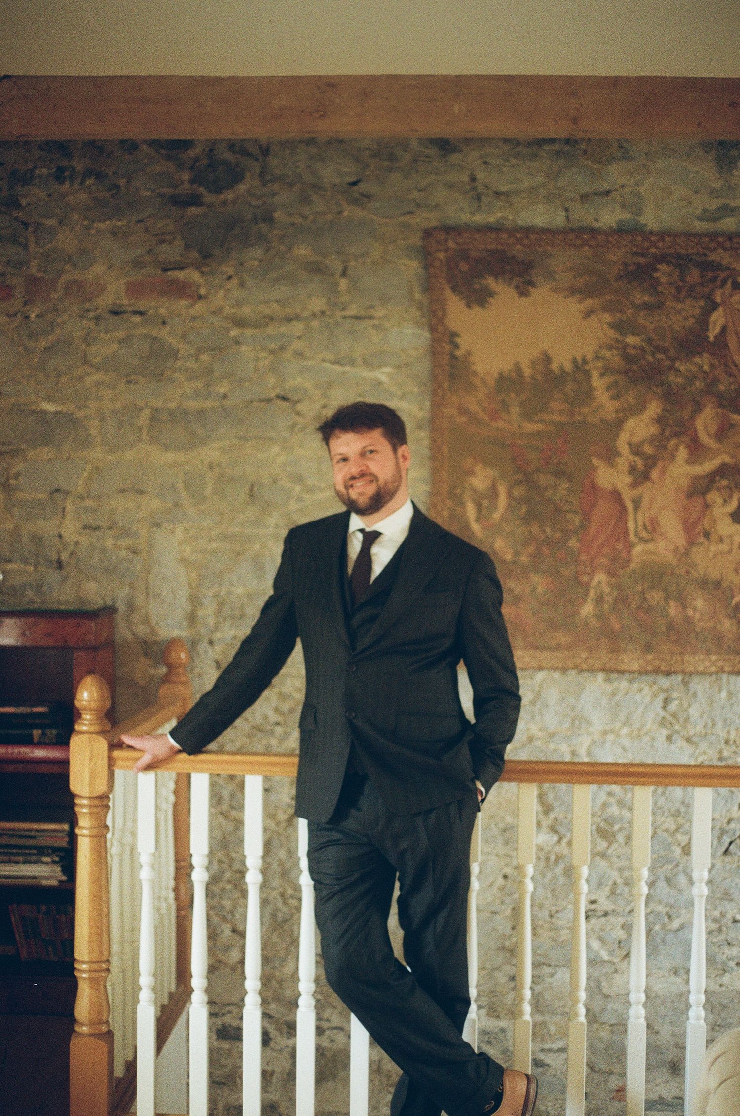 Figure in dark suit poses casually against stone wall with classical artwork and white railing in elegant interior.
