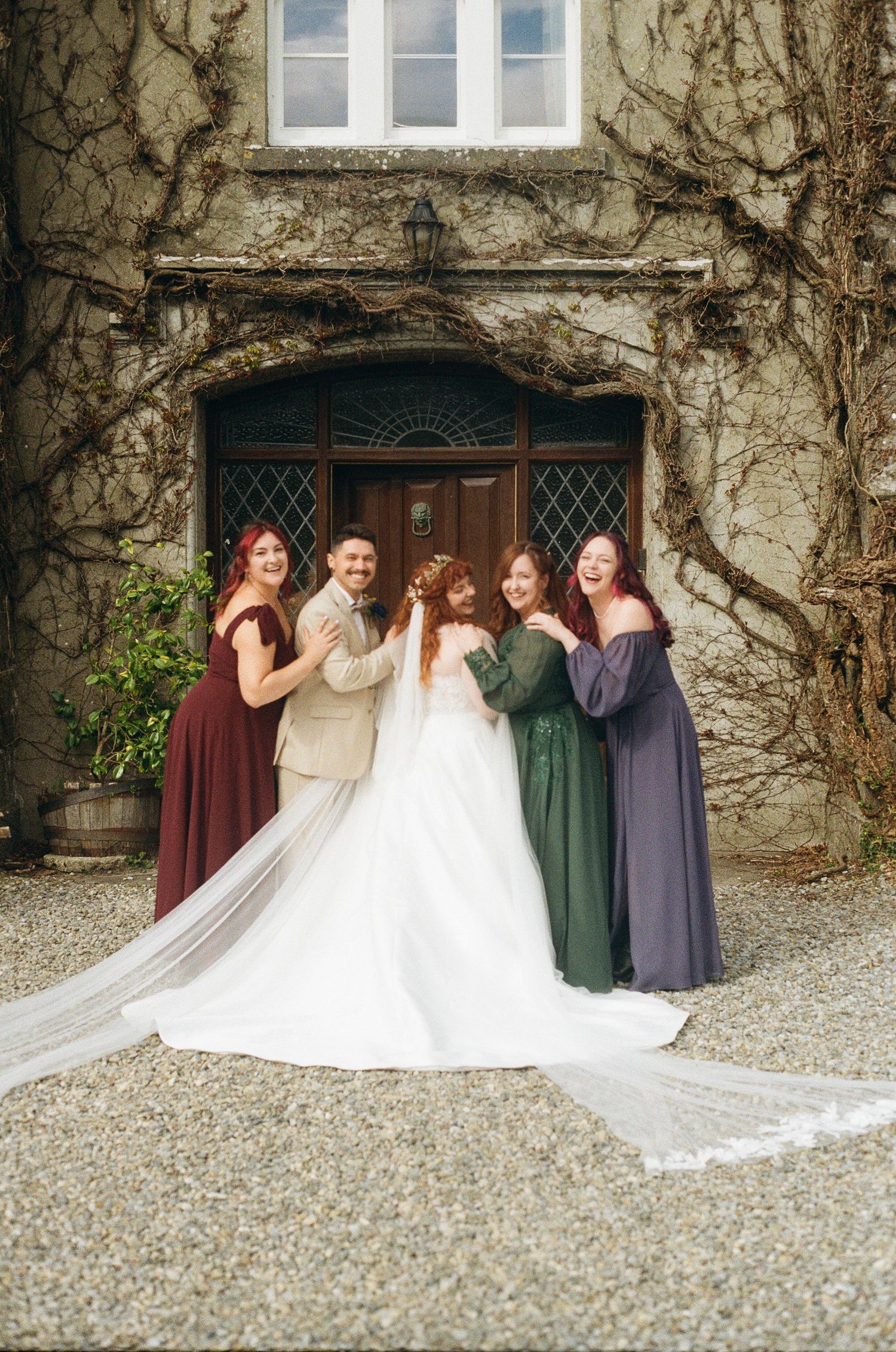 A wedding party poses outside a rustic stone building covered in vines, wearing jewel-toned dresses and formal attire.