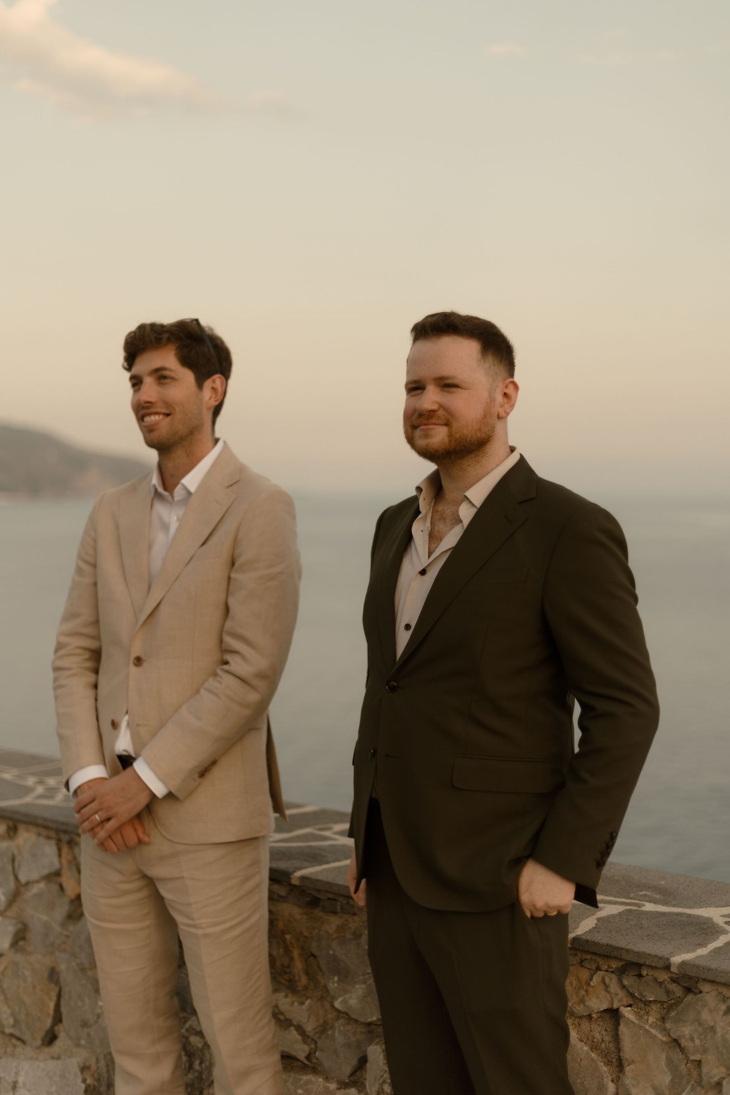 Two people in formal suits stand together on a stone overlook with coastal views at sunset.