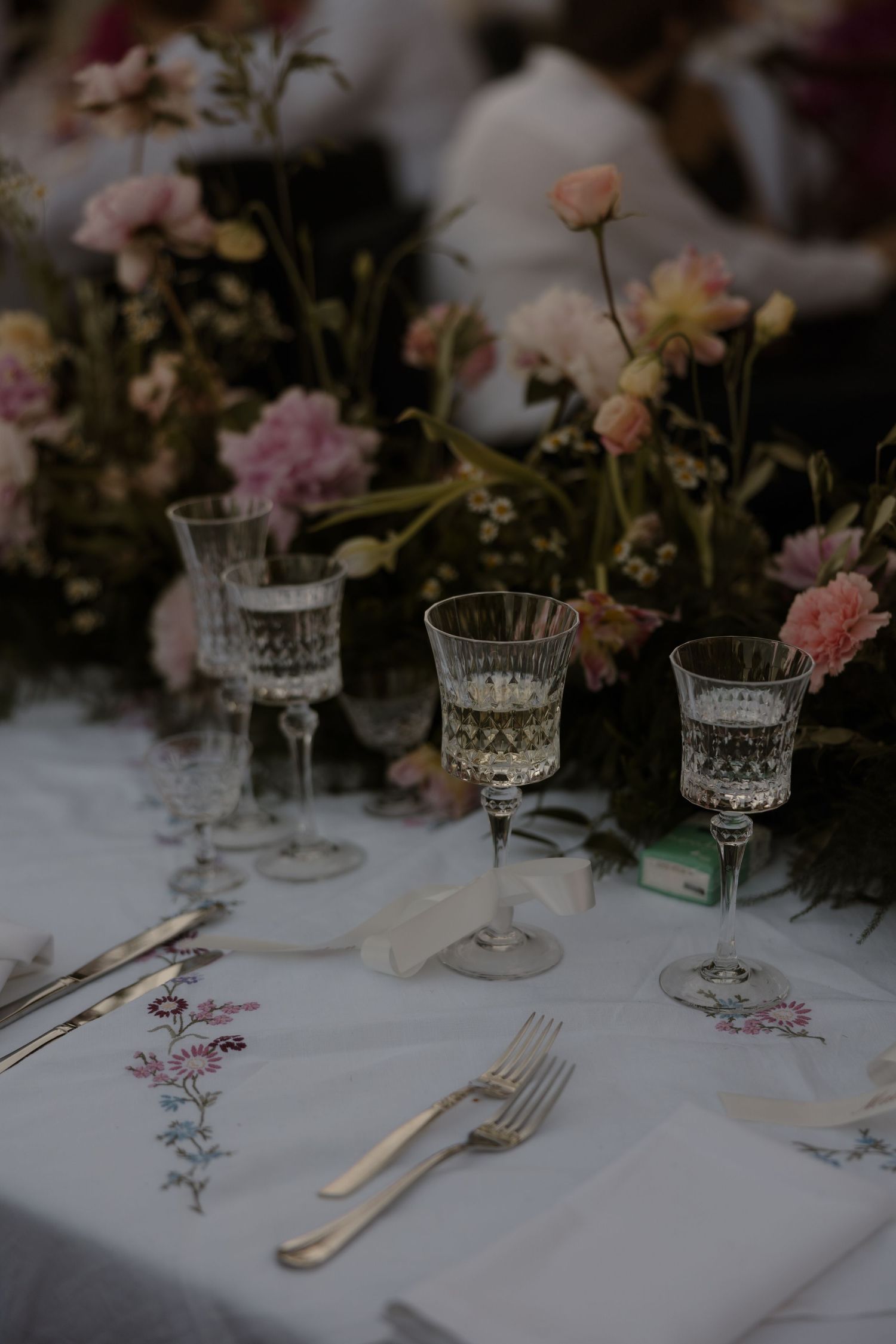 Elegant table setting with crystal wine glasses, delicate flowers, and embroidered linens at romantic outdoor dinner party.
