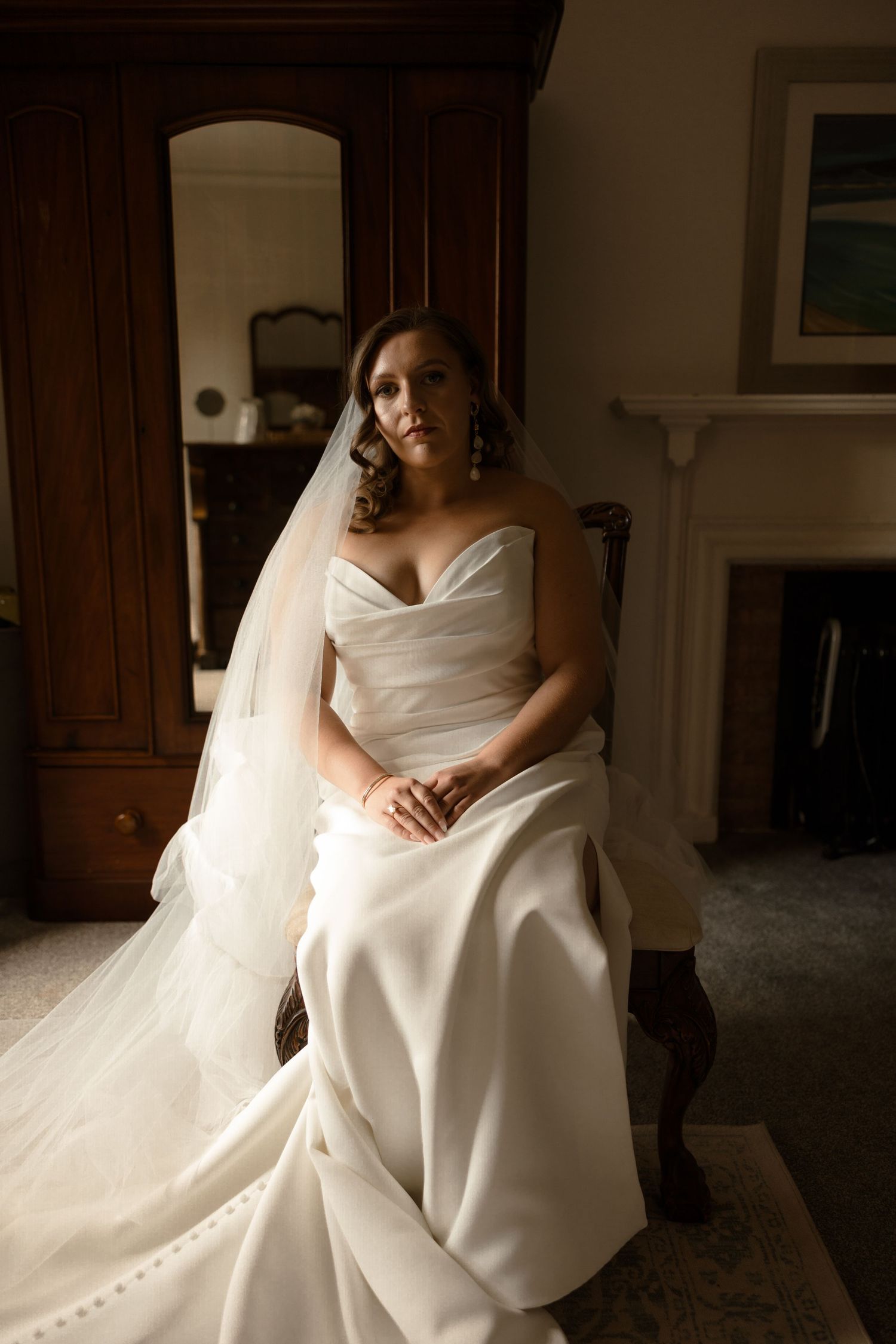 A bride in a sweeping white satin wedding gown and long veil sits elegantly on an antique chair in a romantic interior setting.