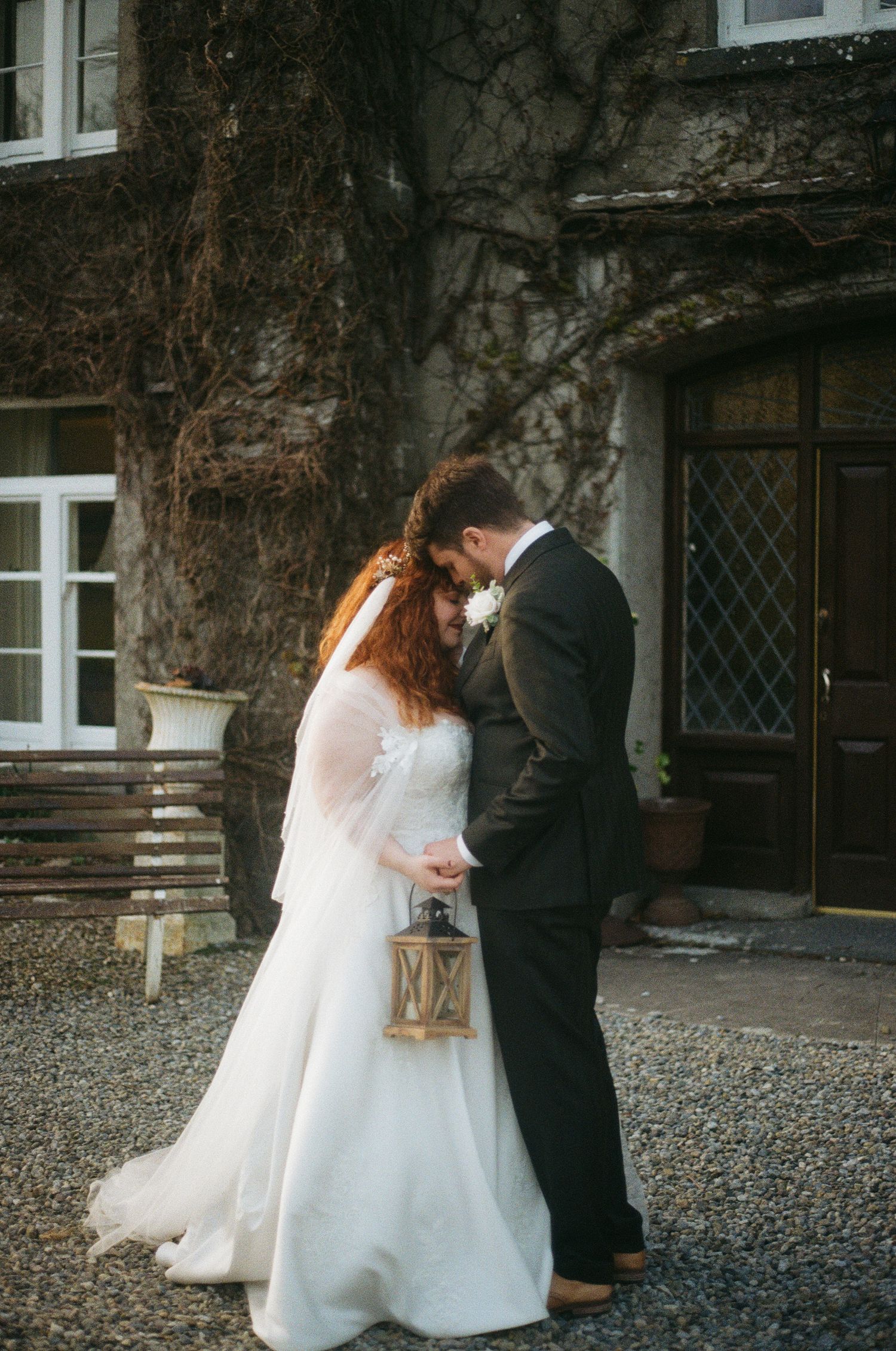A couple shares a romantic moment outside a rustic stone building with climbing vines, the bride holding a vintage lantern.