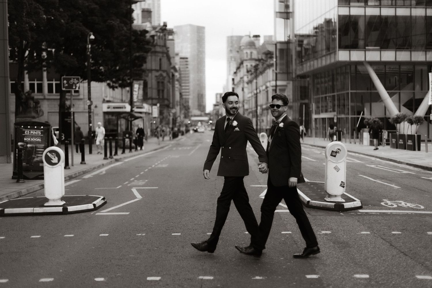 Two people in dark suits walk across a street in a monochrome urban cityscape with buildings in the background.