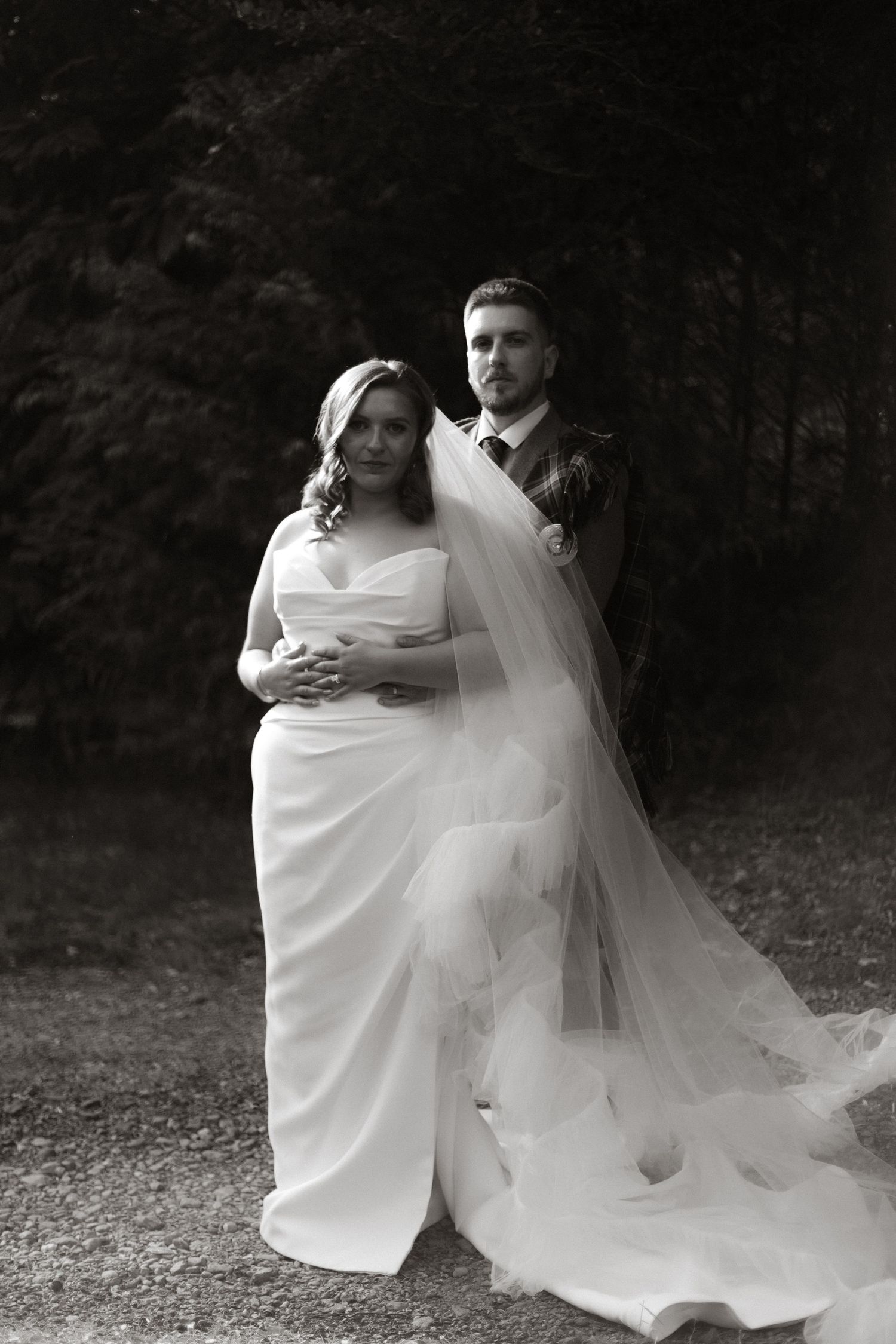 A black and white wedding portrait of a couple outdoors featuring a bride in a flowing gown with a long trailing veil.