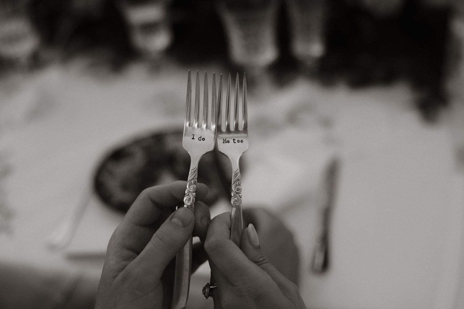 Two hands hold up engraved silver forks against a blurred background in an artistic black and white photo.