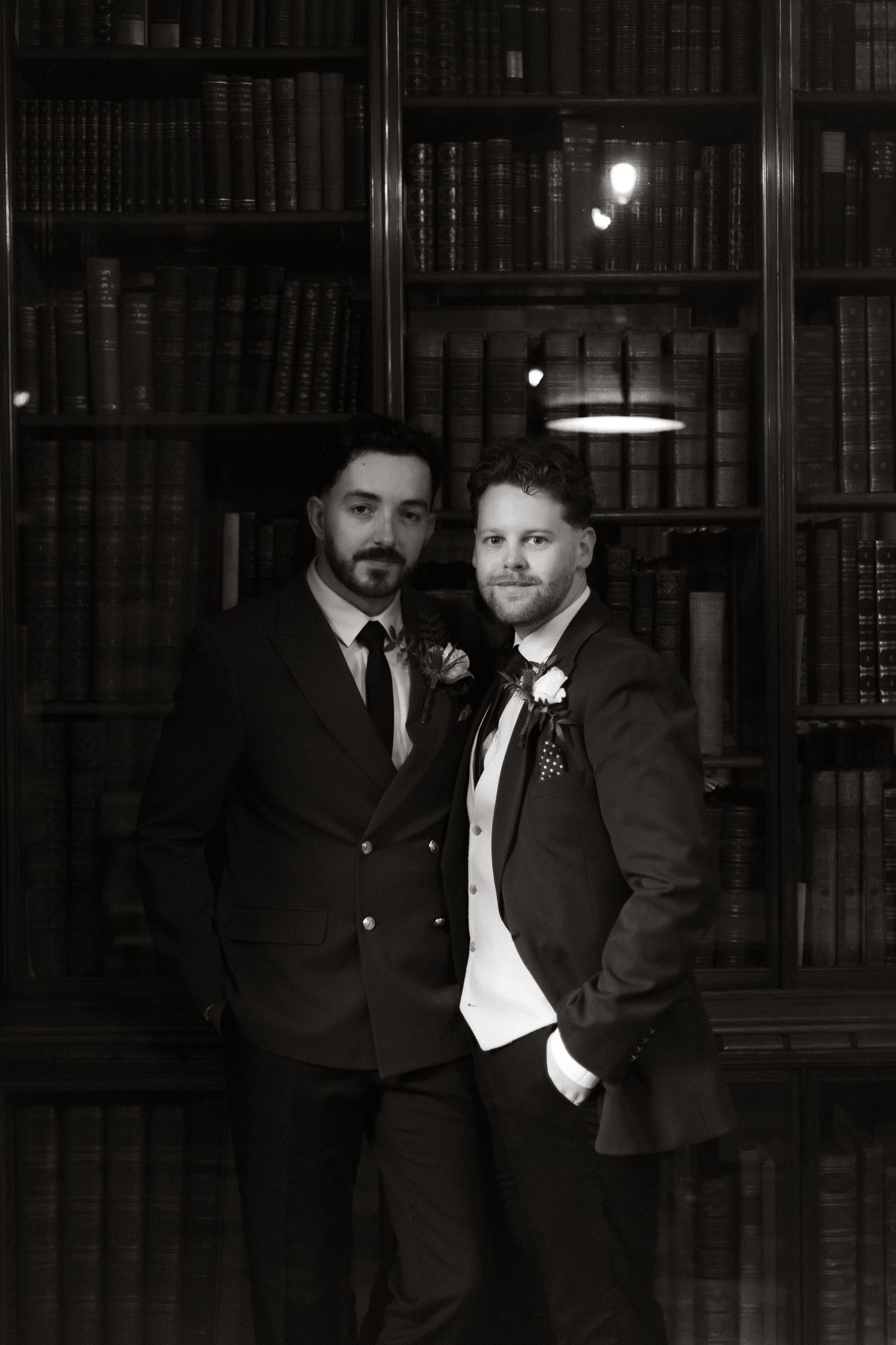 Two people in formal suits pose together in front of a dark library bookshelf with ambient lighting.