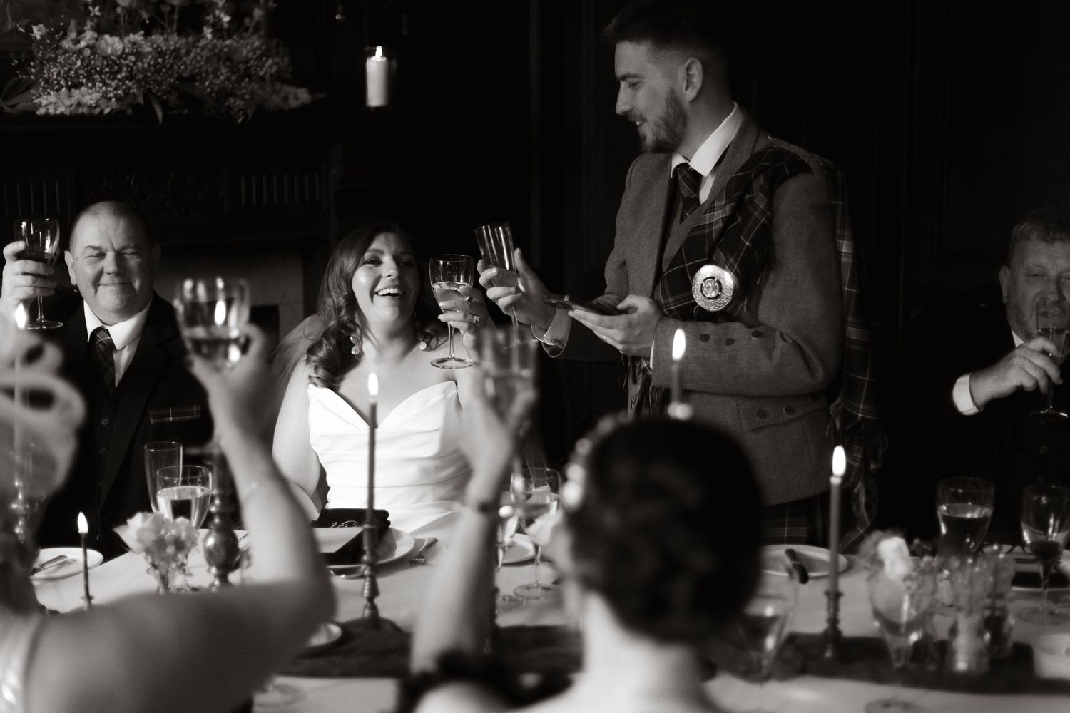 A black and white candid shot of guests raising glasses during a wedding reception toast.