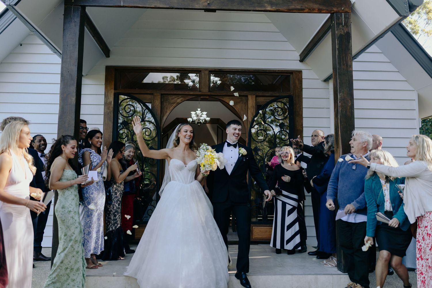 Wedding guests celebrate as newlyweds exit a church building with flower decorations.
