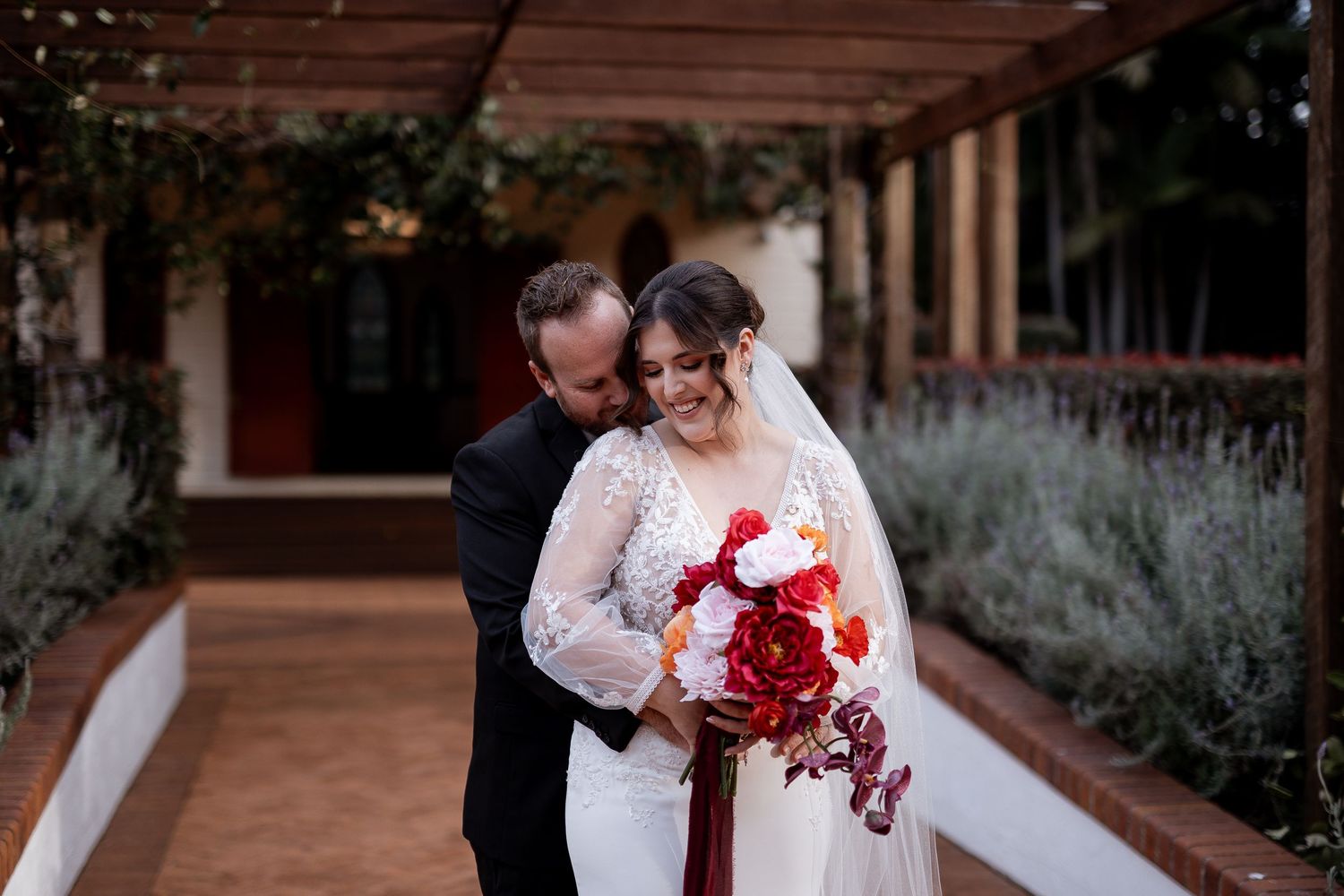 Romantic wedding photo of couple with red rose bouquet under covered walkway at outdoor venue during sunset.