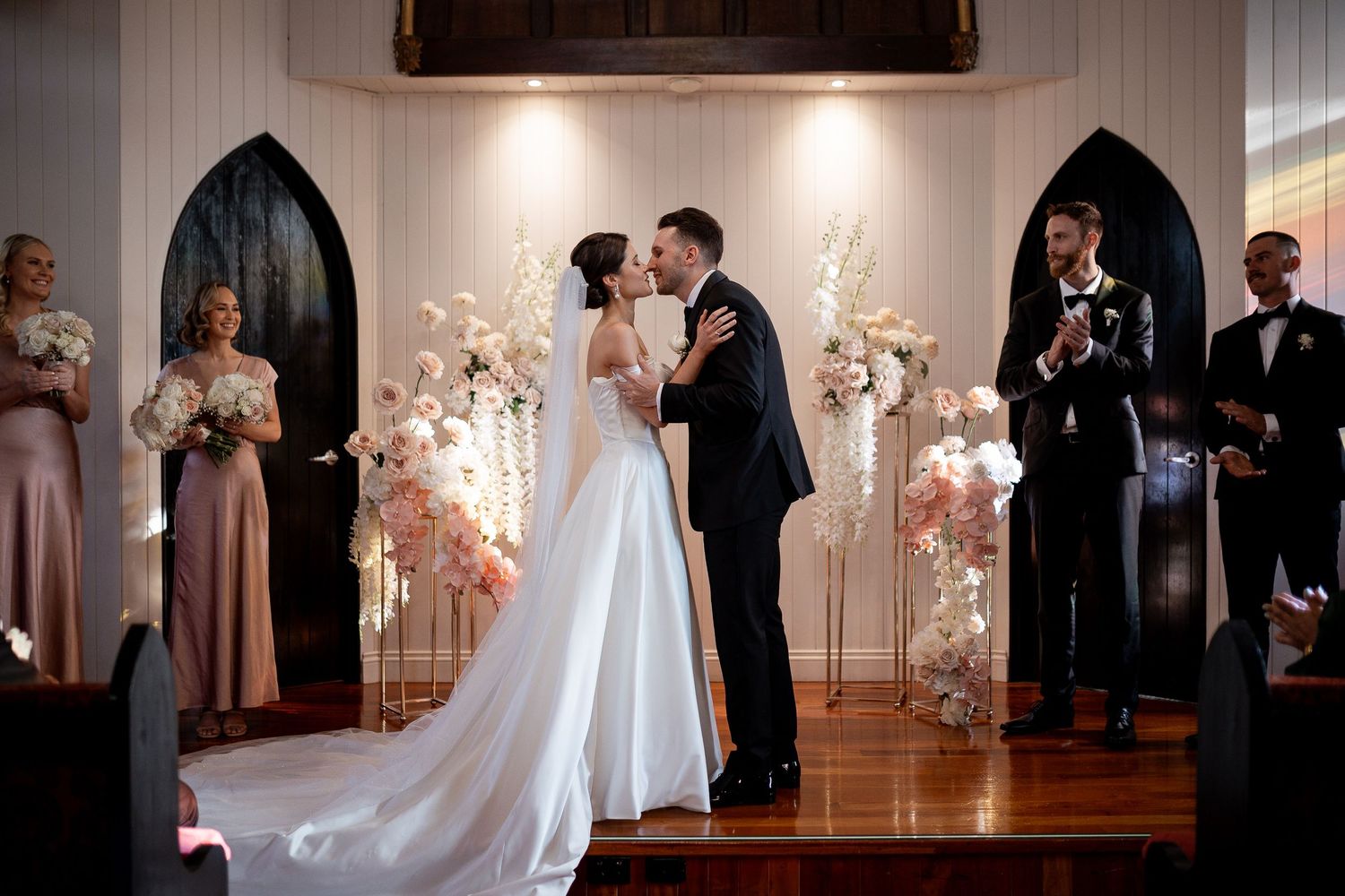 A couple shares their first kiss during a wedding ceremony at an elegant chapel with floral arches and wooden floors.