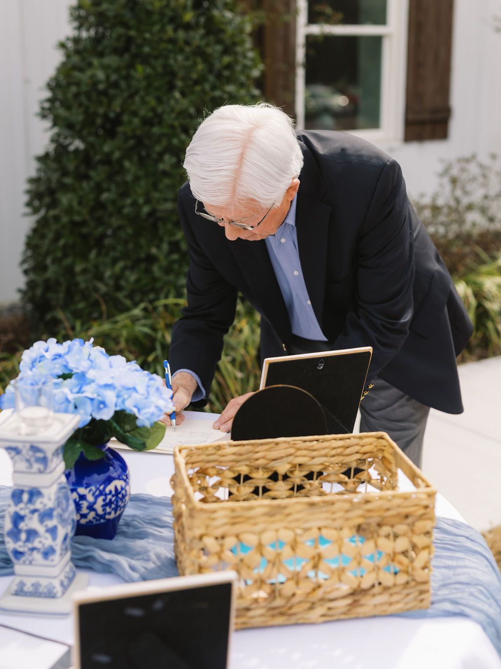 A person in a dark suit leans over a table with a woven basket and blue and white floral arrangement outdoors.
