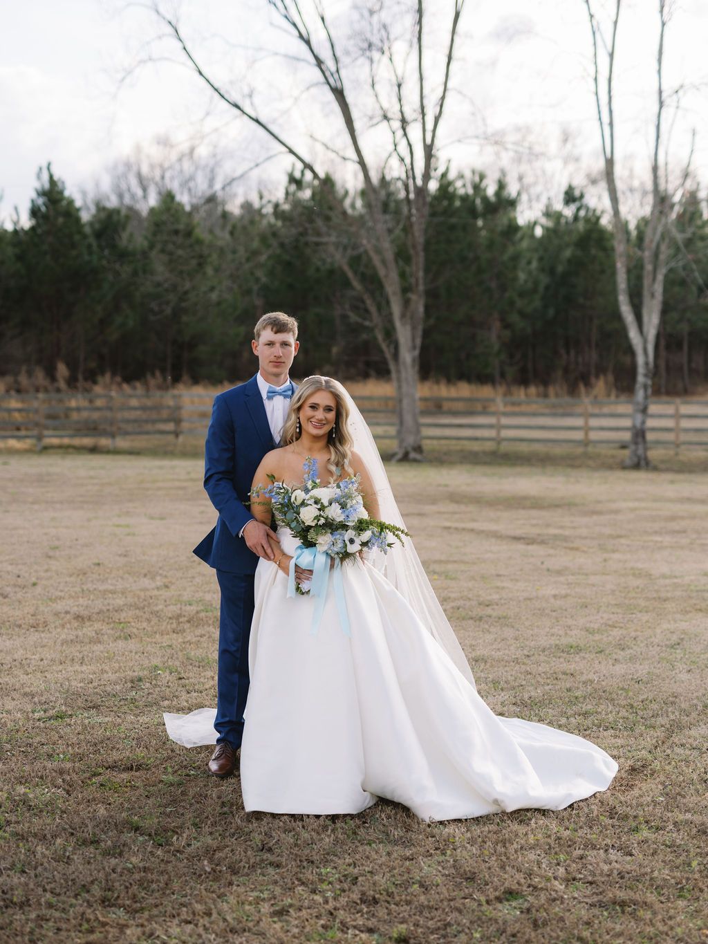 A couple stands together outdoors at their winter wedding, with bare trees and evergreens in the background.
