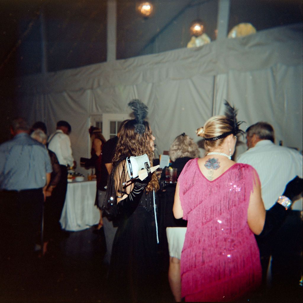 Guests socialize and dance at an evening celebration under a white tent with dim lighting and party decorations.