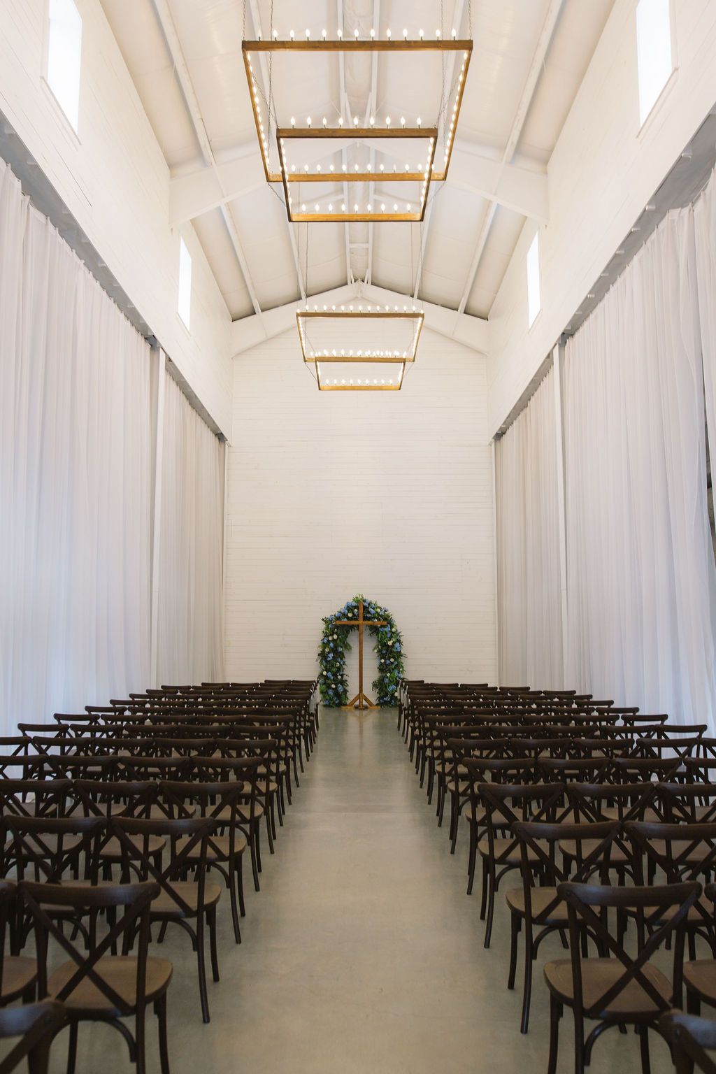 Modern white chapel interior with dark cross-back chairs arranged in rows and elegant brass chandeliers leading to a floral arch.