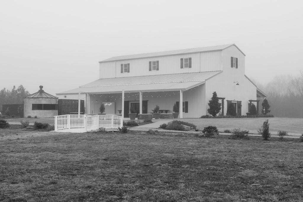 Black and white photograph of a rustic two-story farmhouse with white siding and a wraparound porch in a rural setting.