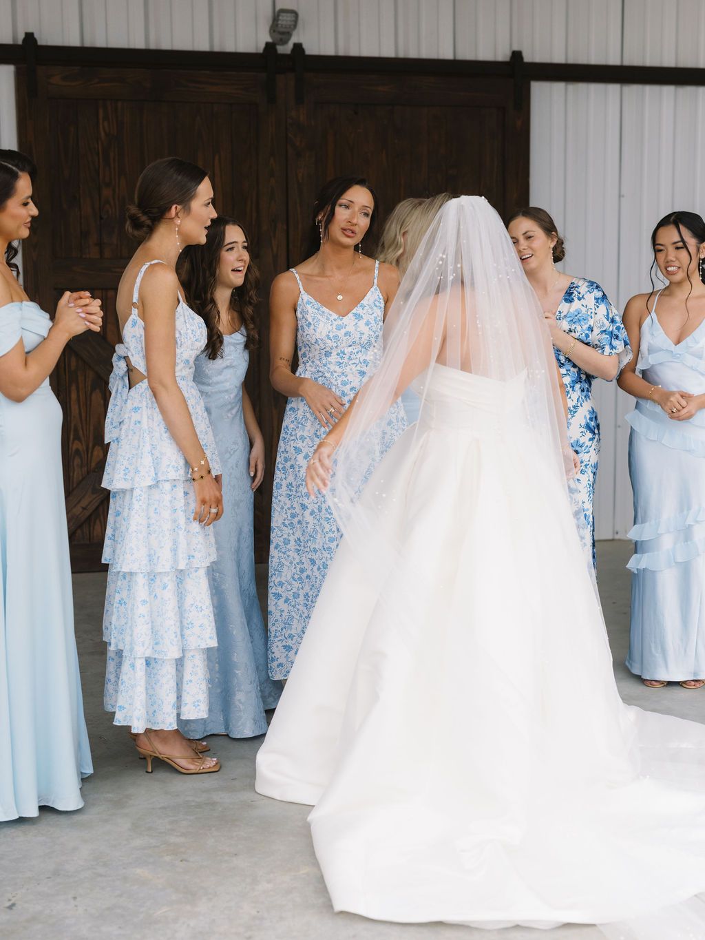 Bridesmaids in light blue floral dresses gather around bride wearing white gown and veil during wedding celebrations.