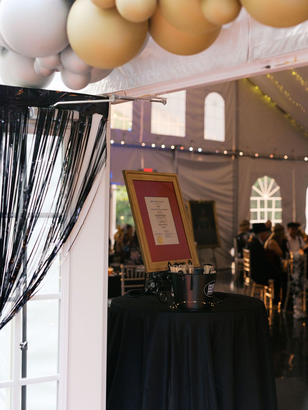 An event venue interior with black curtains, balloons, string lights and a framed sign on a black tablecloth.