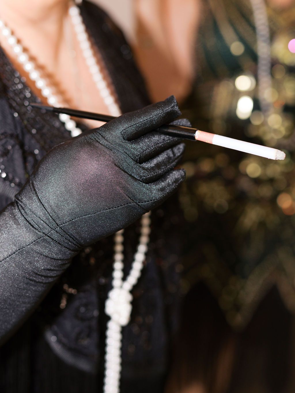 A gloved hand in a black satin evening glove holds a white cigarette against a blurred background with pearl necklaces.