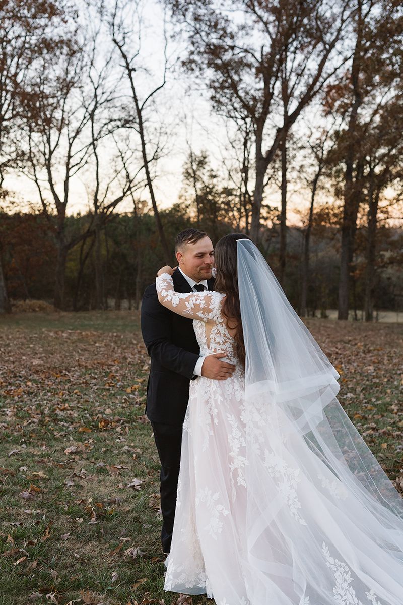 Wedding couple in romantic embrace with dramatic flowing veil at dusk.