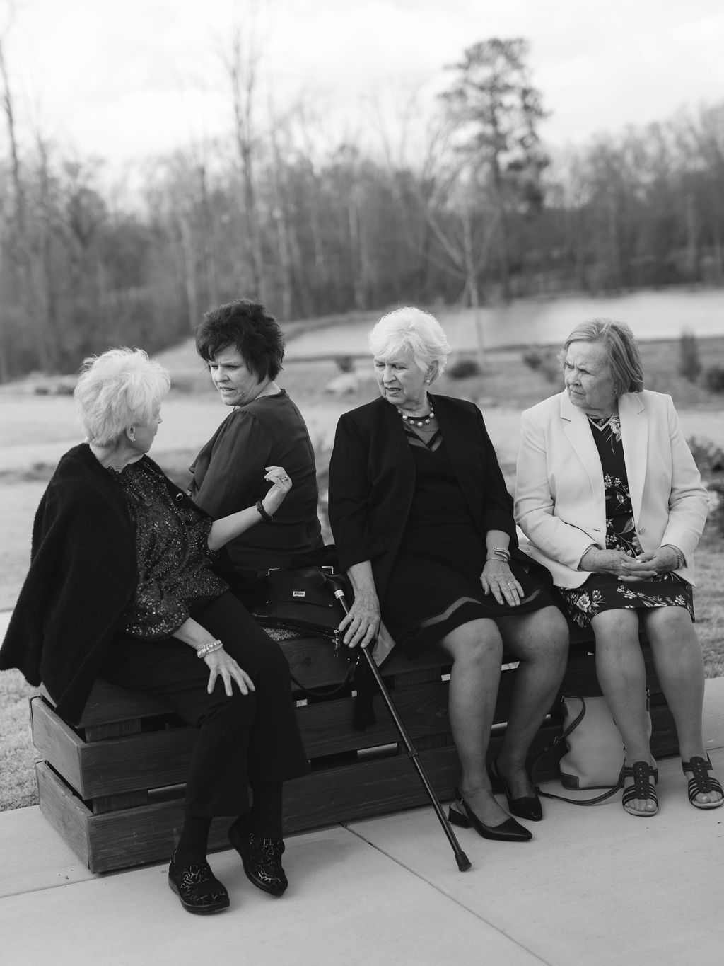 Black and white photograph showing four older women sitting together on a bench outdoors in a park setting.