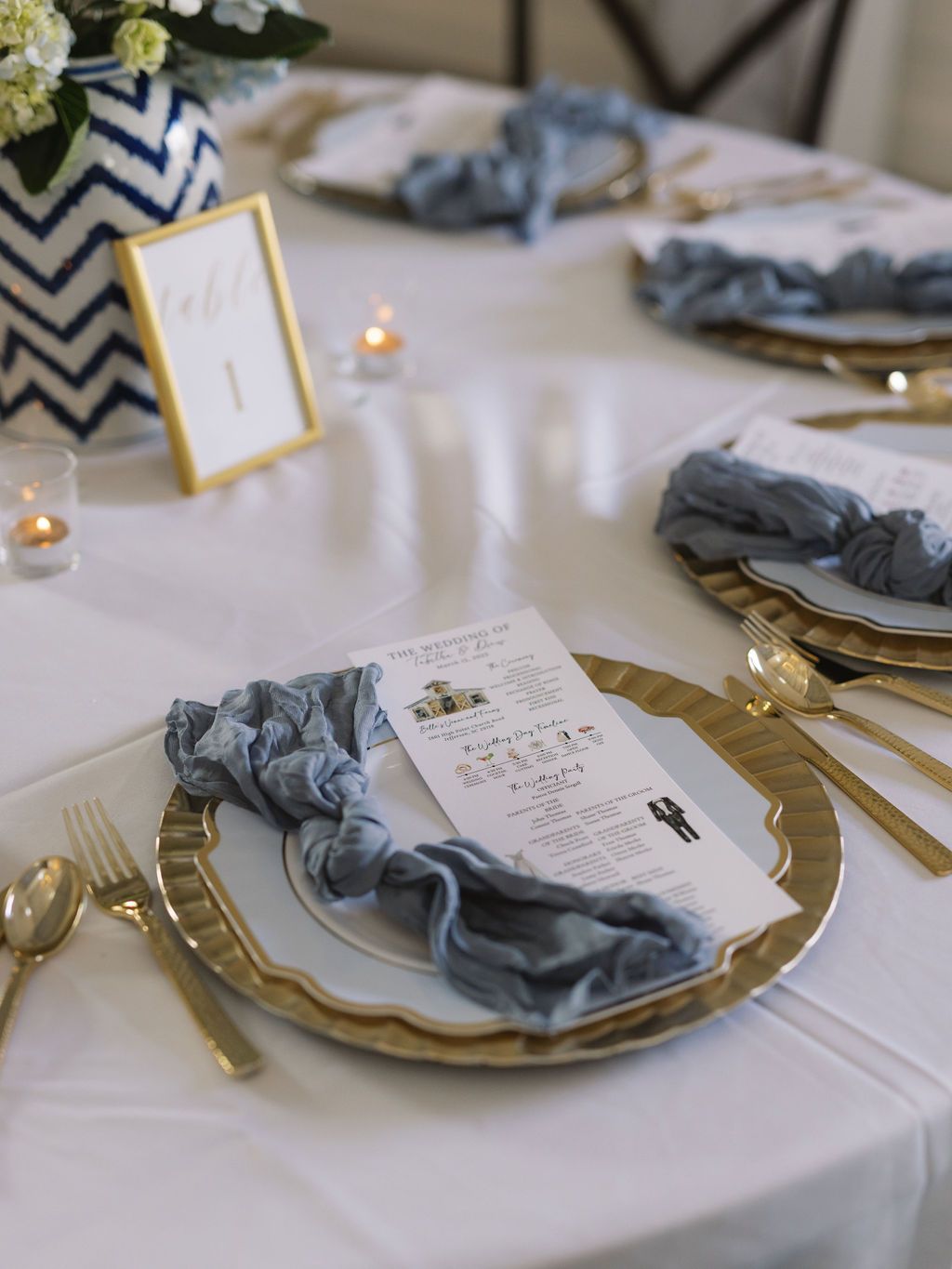 Elegant place setting with gray silk napkin, gold charger plate, and menu card on crisp white tablecloth with soft candlelight.