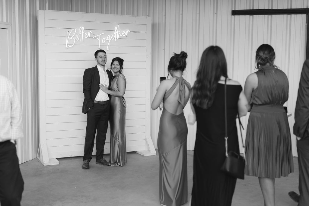 Guests pose for photos at a formal event in front of a white backdrop while others wait their turn in line.