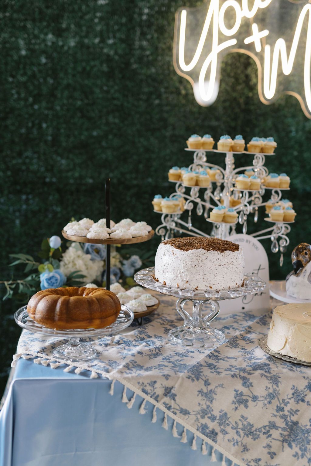 Elegant dessert table with tiered cupcake stands, a bundt cake, and white cake displayed on blue floral tablecloth.