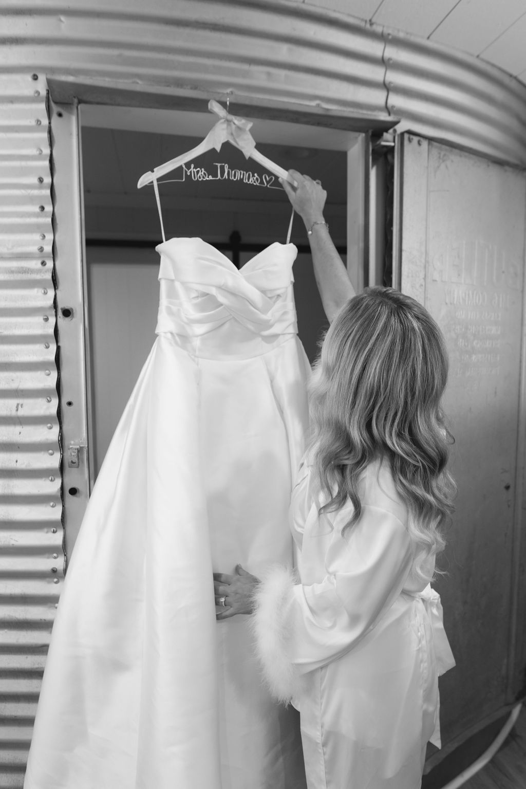 A black and white image shows someone hanging a white wedding dress on a hangar against a curved metal wall.