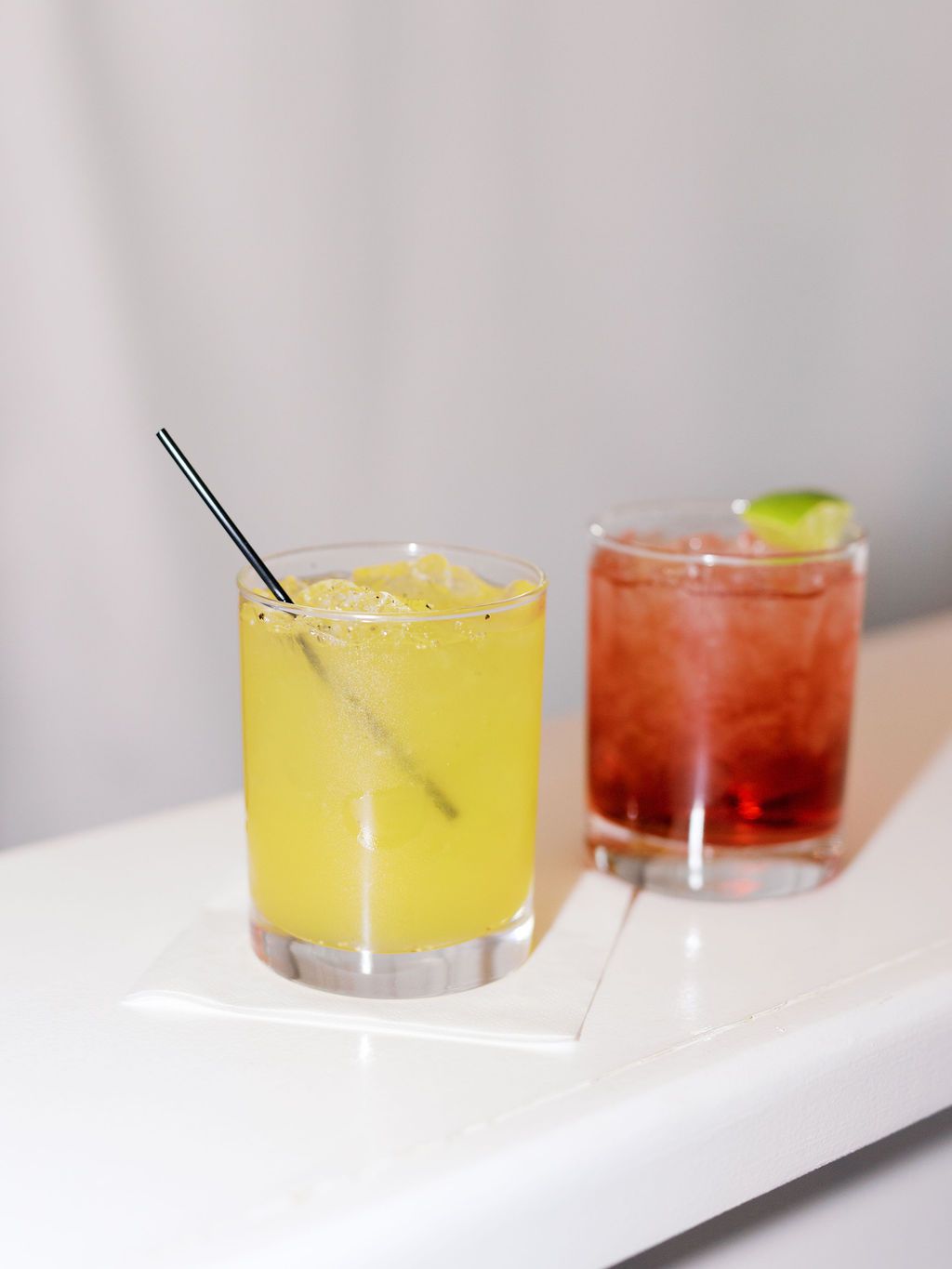 Two craft cocktails on white surface - yellow margarita with salt rim and red drink with lime garnish in rocks glasses.