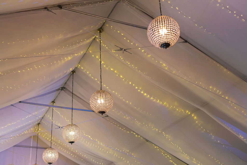 Crystal disco balls and string lights illuminate a white tent ceiling creating an elegant party atmosphere.