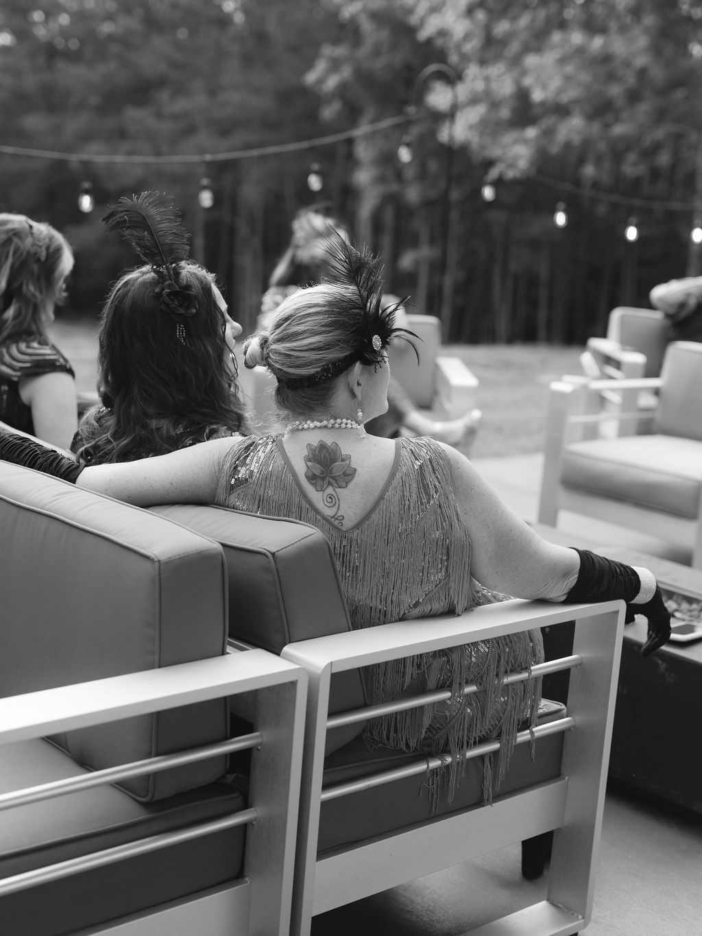 People sit outdoors at a casual evening event with string lights glowing overhead in black and white.