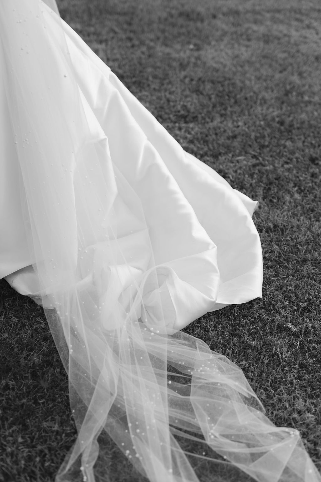 Black and white photograph of flowing wedding dress veil laying on grass with delicate fabric details.