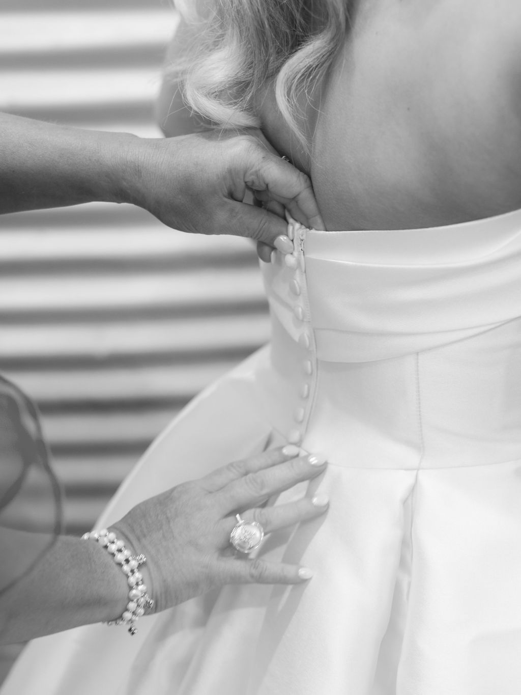 Close-up black and white photo of hands buttoning up a white wedding dress with pearl bracelet and engagement ring visible.
