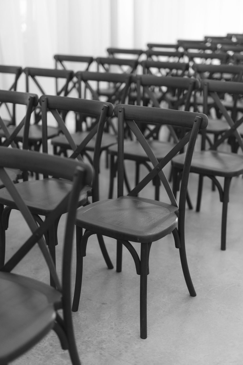 Empty black wooden cross-back chairs arranged in rows on a concrete floor, shown in black and white photography.