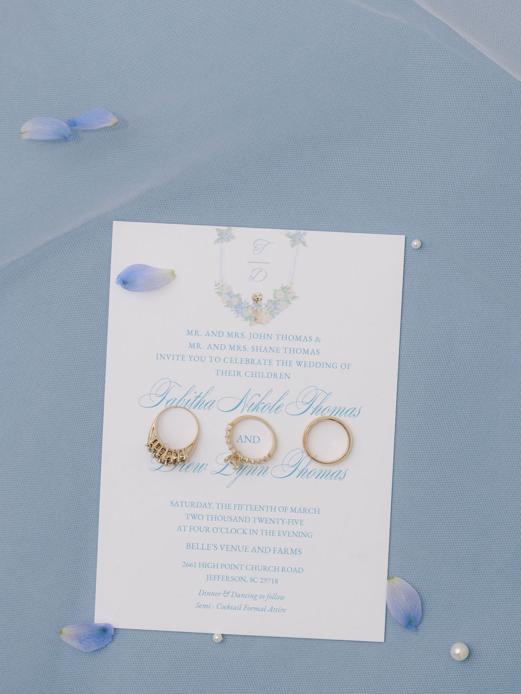 Elegant wedding invitation with three gold rings displayed on a light blue surface with scattered purple flower petals.