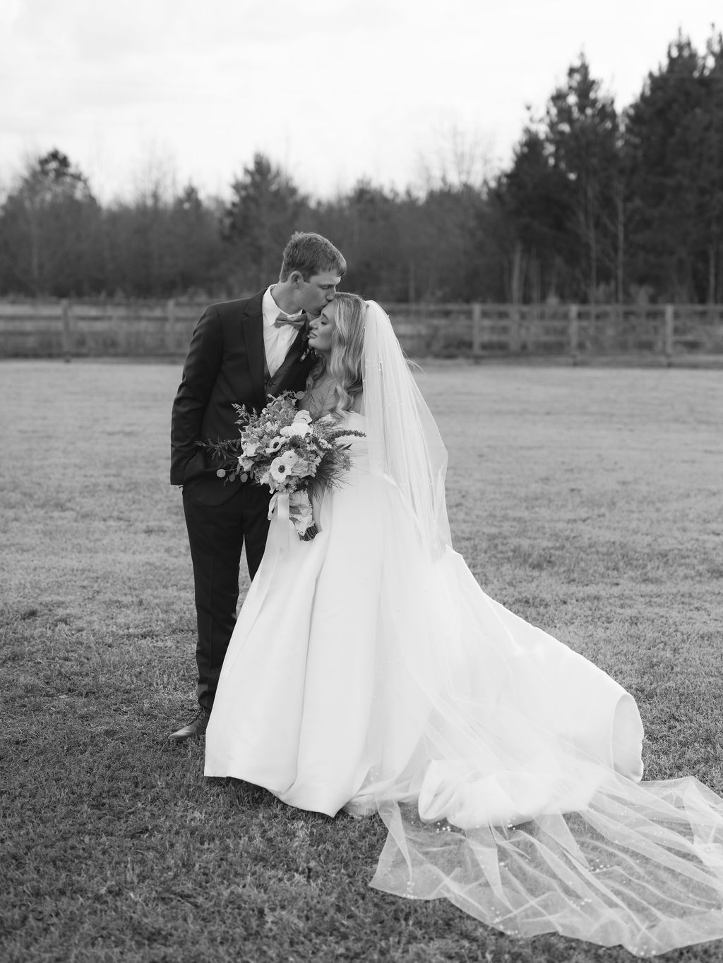 A romantic black and white wedding photo of a couple sharing a tender moment in an open field with pine trees in background.