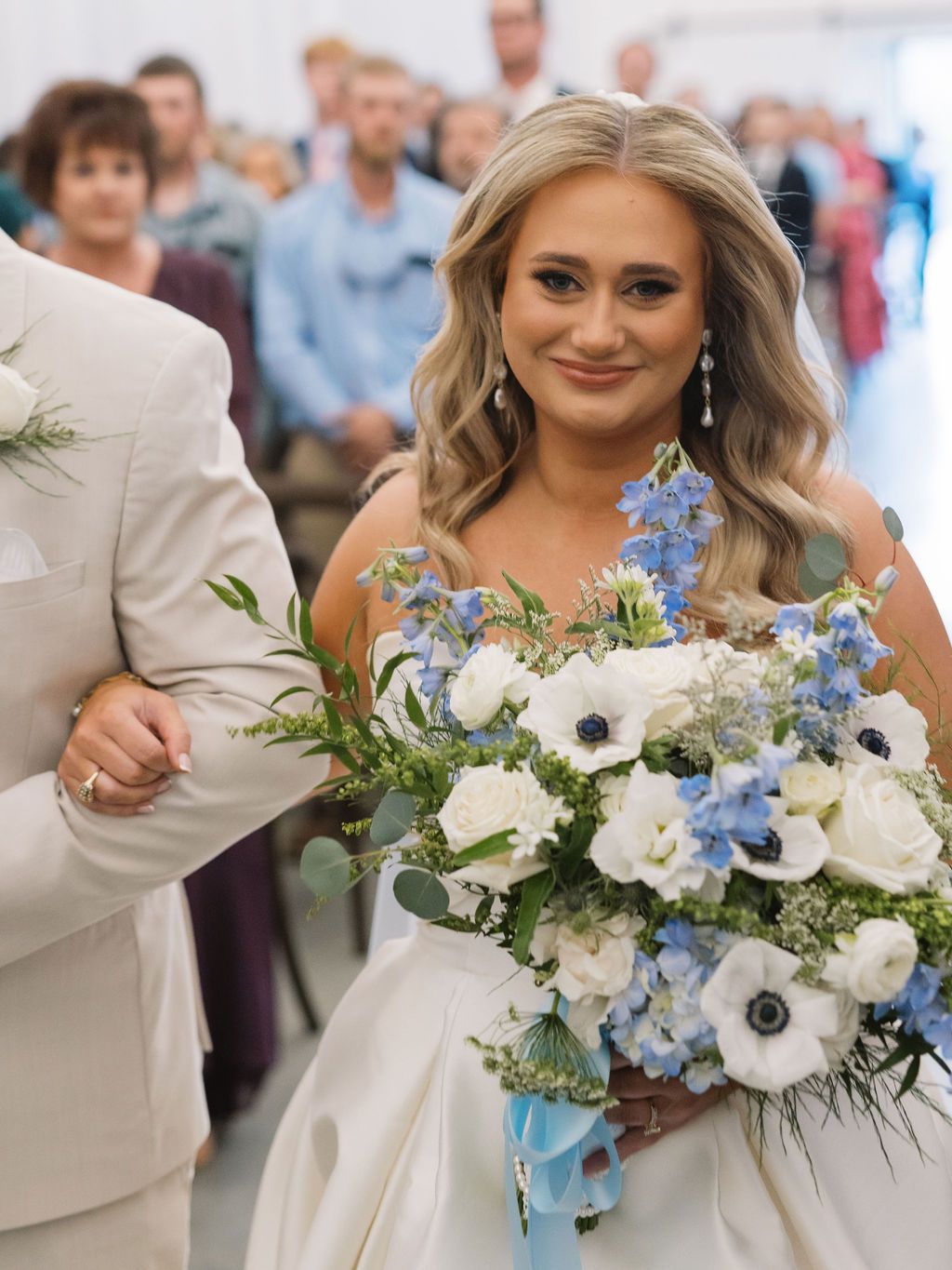 A bride wearing a strapless white gown holds a stunning bouquet of white anemones and blue delphinium flowers.