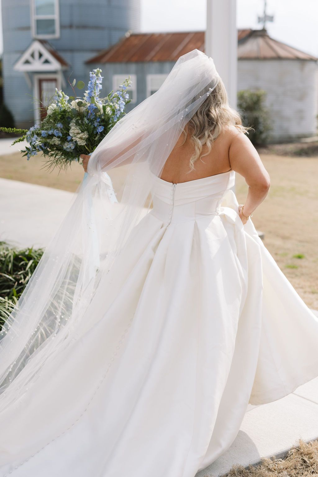 Back view of a bride in a flowing white wedding gown and veil holding a blue and white floral bouquet outdoors.