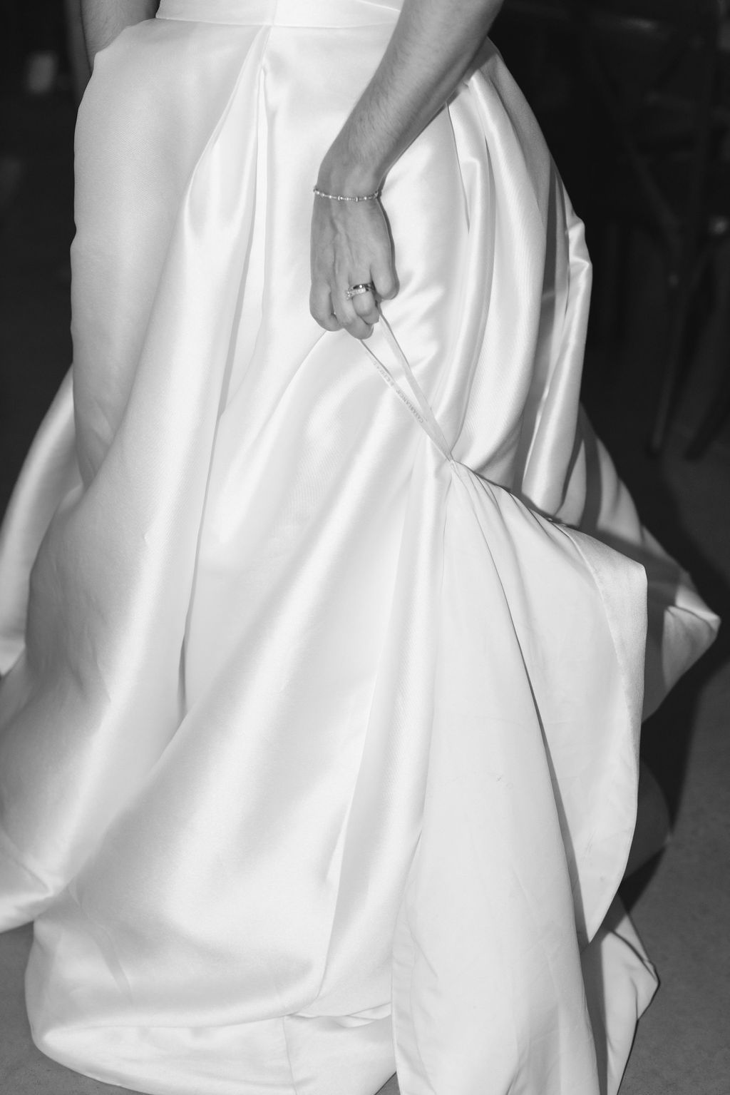Black and white close-up of hands holding gathered fabric of an elegant white wedding dress, showing luxurious pleats and folds.