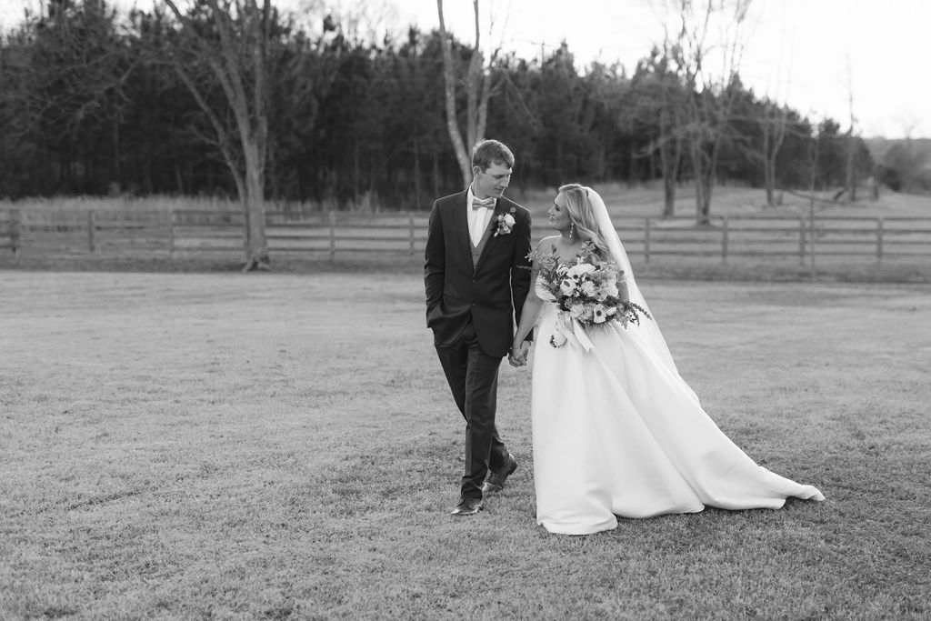 A couple in wedding attire walk together on a grassy lawn with trees in the background captured in black and white.