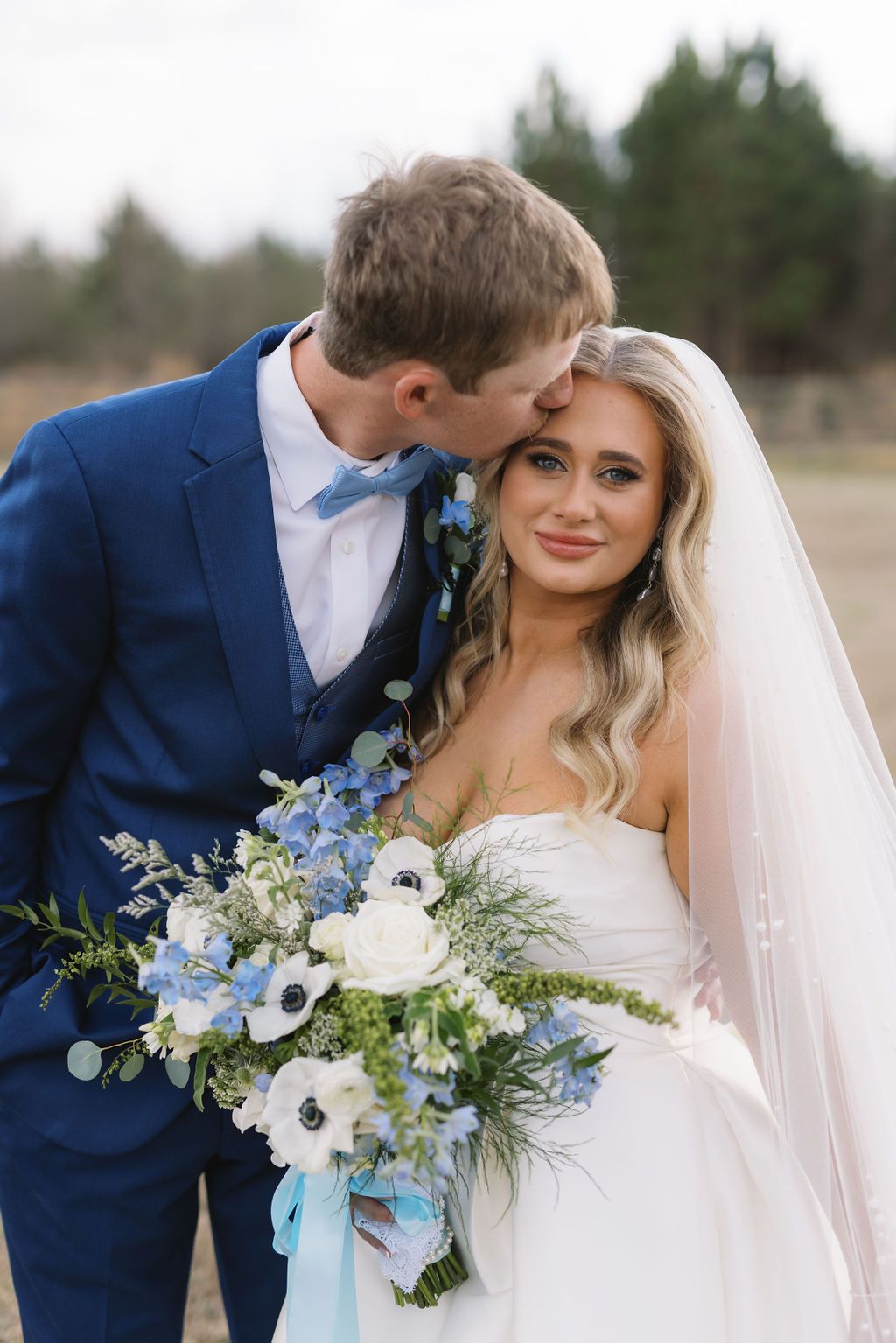 A couple shares a tender moment on their wedding day, with a striking blue and white floral bouquet against a navy suit.