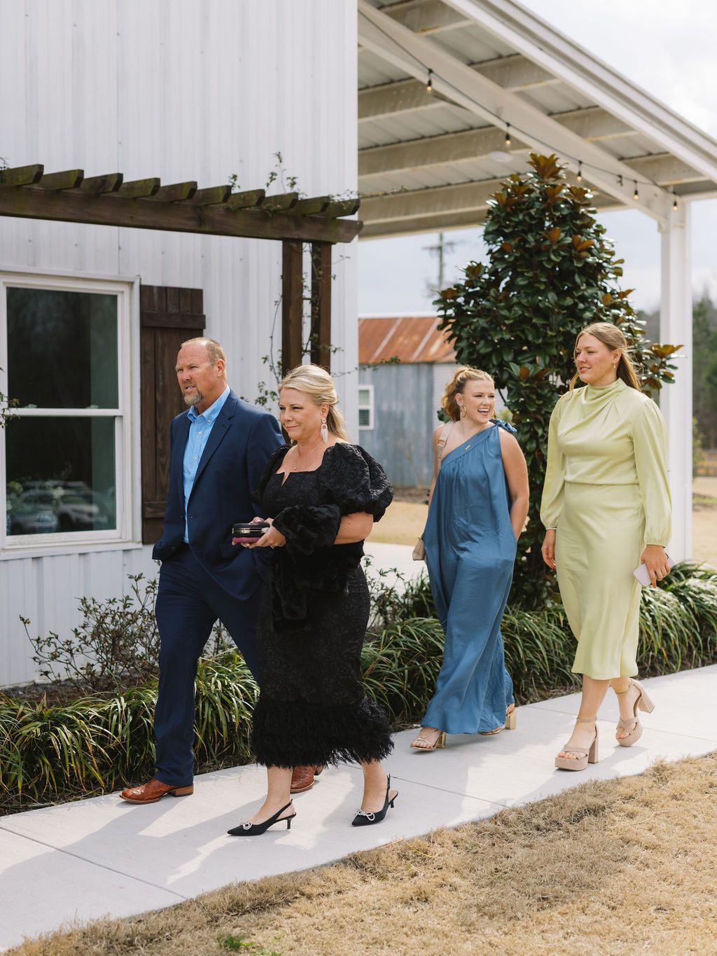 A formal gathering with people walking in a line outside a white building with a pergola and plants.