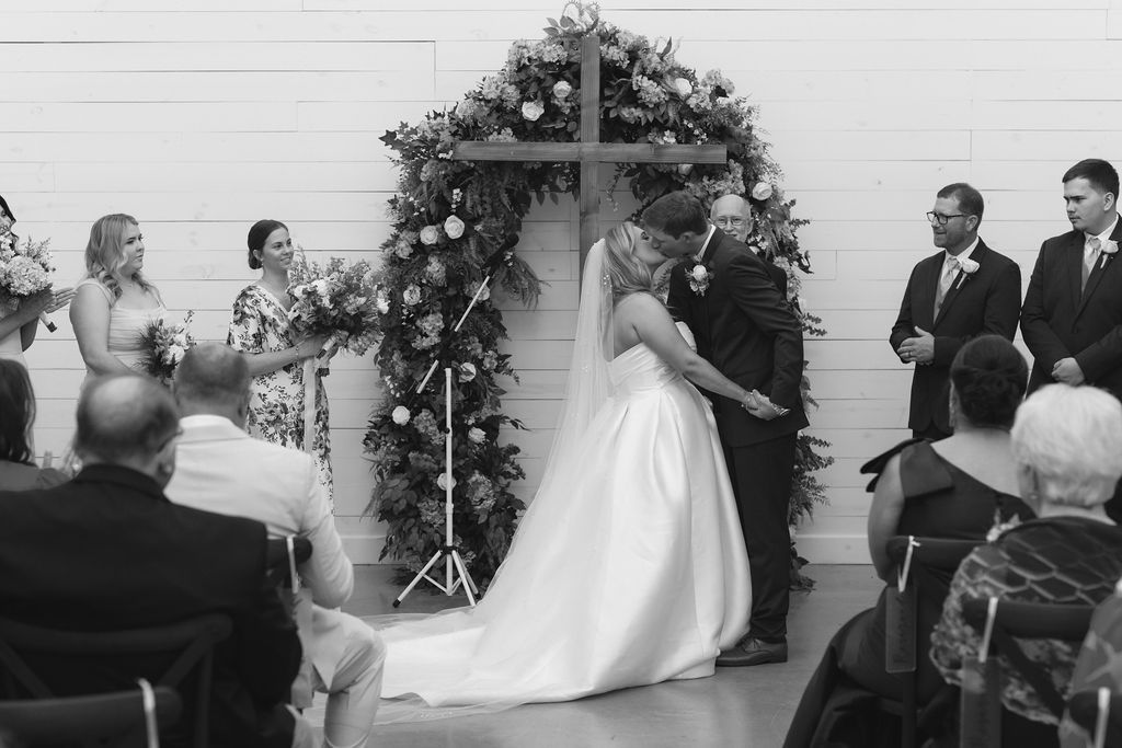 Black and white photo of a wedding ceremony taking place under a floral arch with guests seated in attendance.