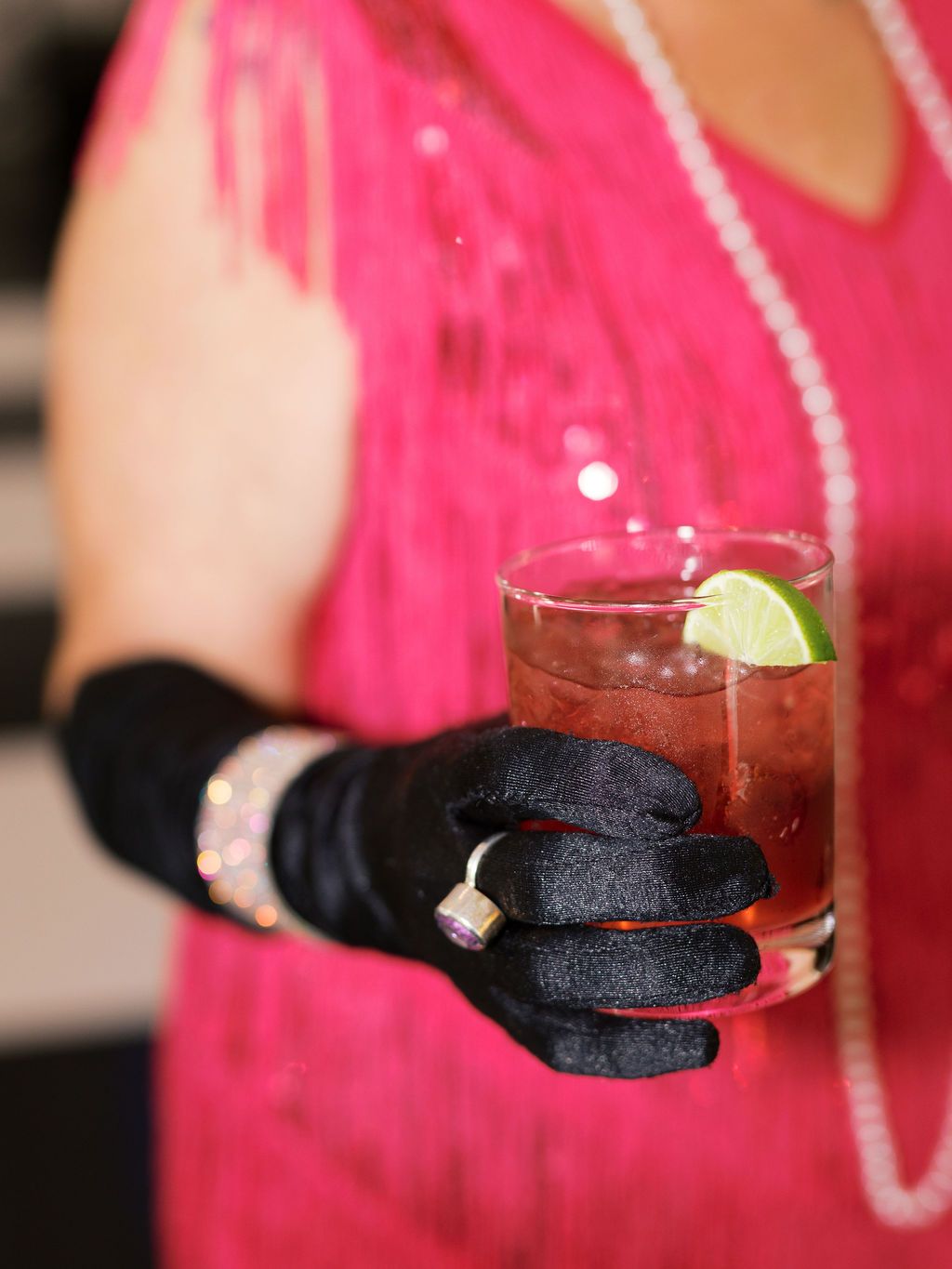 A gloved hand holds a cocktail with a lime garnish against a pink dress background.