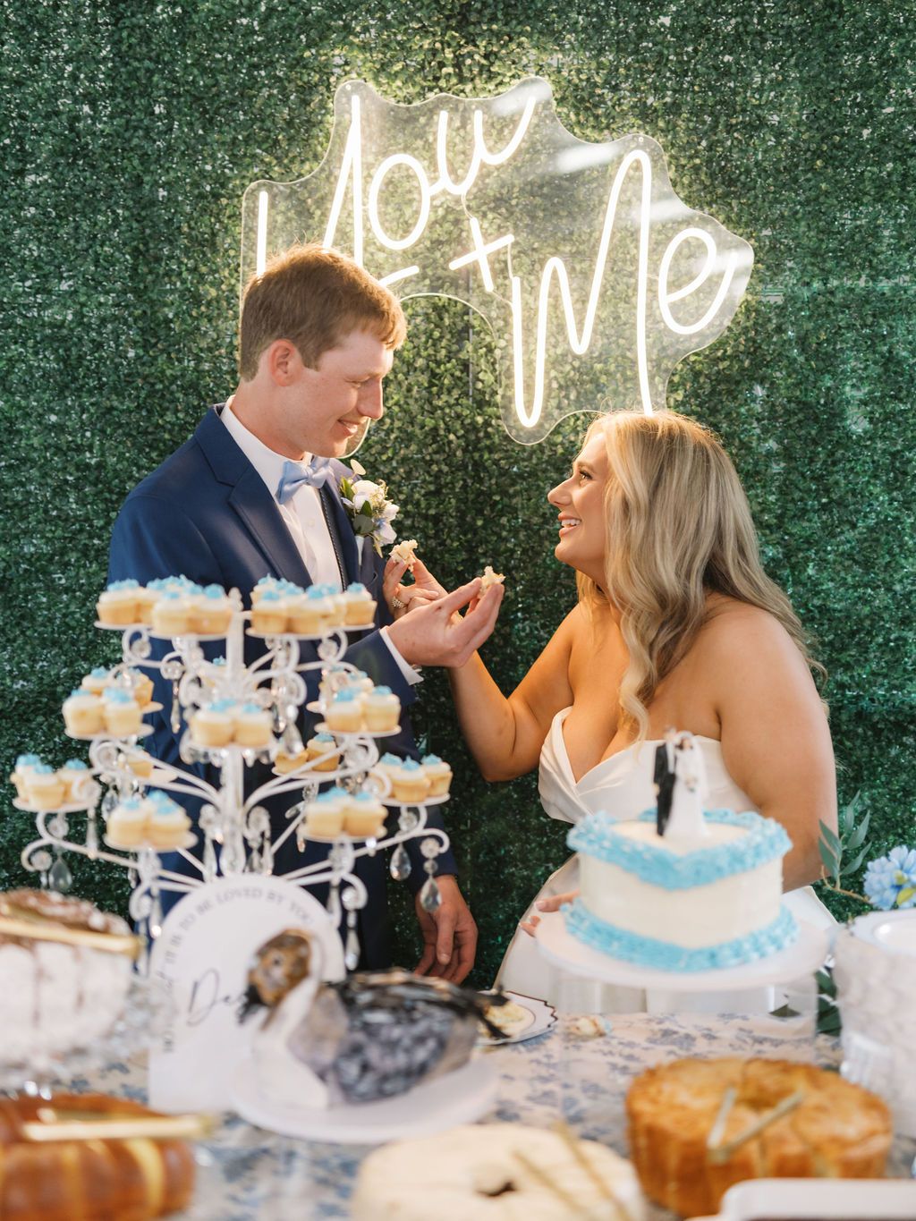 Newlyweds share a sweet moment at their wedding dessert table with cupcakes, cake and a neon love sign against greenery.