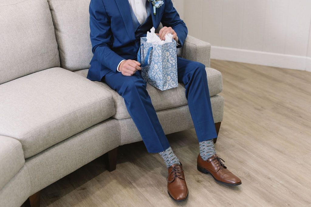 Figure in blue suit sits on beige sofa holding wrapped gift, brown dress shoes on wooden floor.